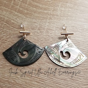 May include: A pair of fan-shaped earrings. One is dark grey with carved patterns, the other is iridescent white with similar carvings. Both have gold-coloured posts and bars. The earrings are displayed on a wooden surface.