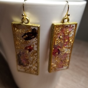 May include: A pair of gold-coloured rectangular earrings. Each earring has clear resin filled with gold leaf and small pieces of dried flowers in red and pink. The earrings have a simple hook closure.