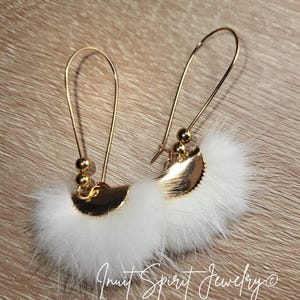May include: Gold-coloured drop earrings with white fluffy detailing. The earrings feature a curved wire design and small gold bead accents. The earrings are displayed on a wooden surface. The text "Inuit Spirit Jewelry" is visible.