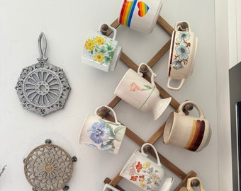 May include: A wooden mug rack with 10 mugs hanging on it. The mugs are all different colors and patterns, including floral, rainbow, and stripes. The rack is mounted on a white wall.
