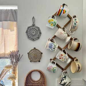 May include: A wooden mug rack with a variety of colorful mugs with floral and geometric designs. The rack is mounted on a white wall with a wicker basket and two trivets.