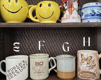 May include: A shelf with eight ceramic mugs. The mugs are decorated with various designs, including smiley faces, cats, a windmill, a globe, a butterfly, and the words "World's Greatest Grandma".