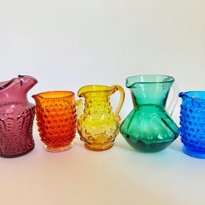 May include: Five colorful glass pitchers in shades of purple, orange, yellow, green, and blue. Each pitcher has a unique shape and design, with textured surfaces and handles. The pitchers are arranged in a row against a white background.