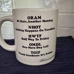 May include: White ceramic coffee mug with black text that reads "ORAM Oh Rats, Another Monday NHOT Nothing Happens On Tuesdays HWTF Half Way To Friday OMDL One More Day Left TGIF Thank Goodness It's Friday".