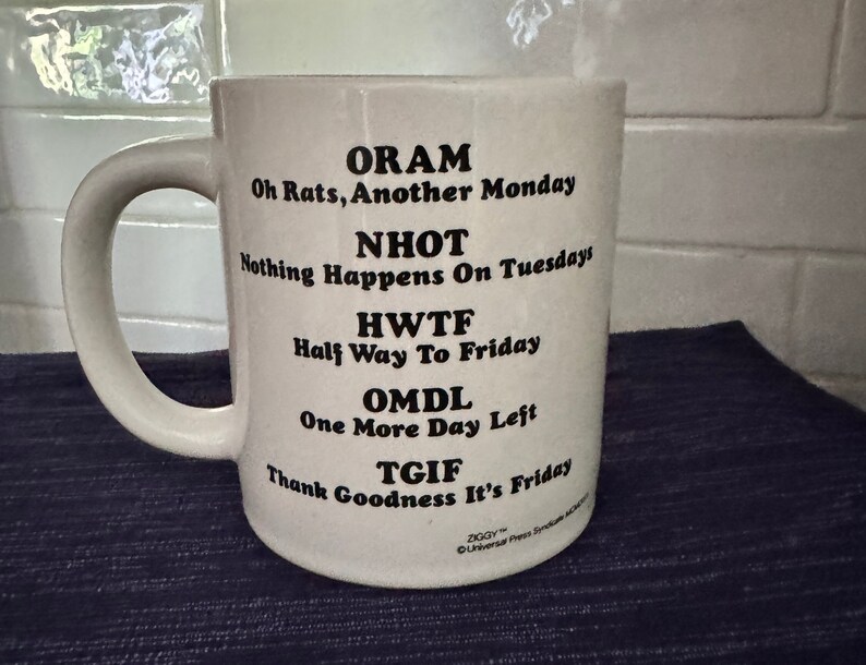 May include: White ceramic coffee mug with black text that reads "ORAM Oh Rats, Another Monday NHOT Nothing Happens On Tuesdays HWTF Half Way To Friday OMDL One More Day Left TGIF Thank Goodness It's Friday".
