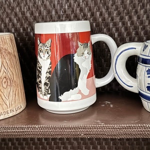 May include: Three ceramic mugs with different designs. The first mug is brown and has a wood grain pattern. The second mug is white with a red background and a black and white cat design. The third mug is white with a blue and white design that resembles a barrel.