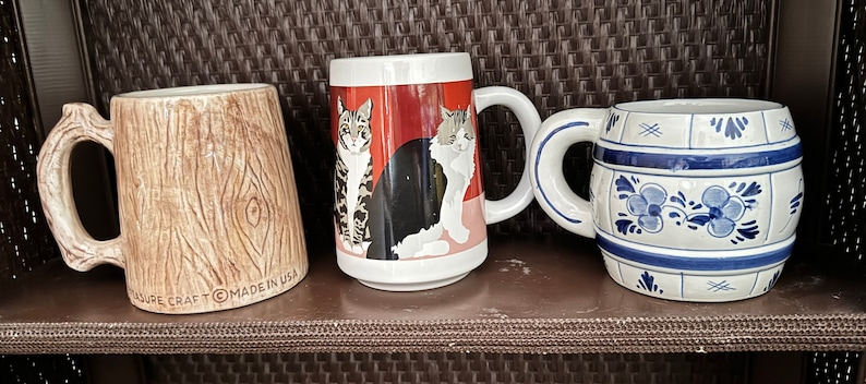 May include: Three ceramic mugs with different designs. The first mug is brown and has a wood grain pattern. The second mug is white with a red background and a black and white cat design. The third mug is white with a blue and white design that resembles a barrel.