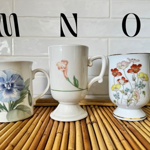 May include: Three white ceramic mugs with floral designs. The mugs have handles and are sitting on a woven bamboo mat. The mugs are labeled M, N, and O.