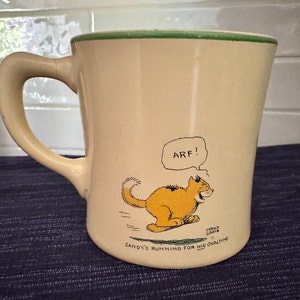May include: A cream-colored ceramic mug with a green rim features a cartoon illustration of a cat running with the words "Arf!" and "Sandy's running for his Ovaltine" printed on the mug.