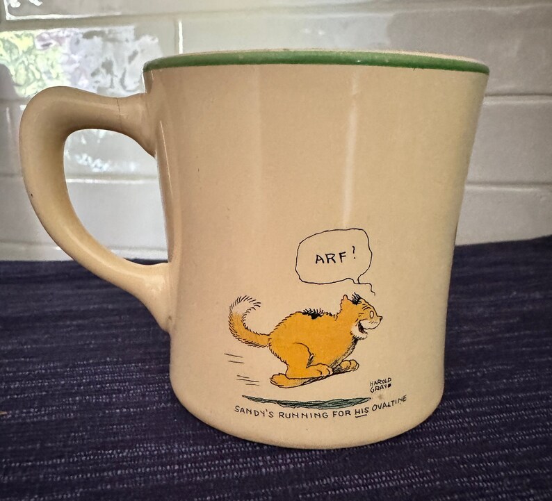 May include: A cream-colored ceramic mug with a green rim features a cartoon illustration of a cat running with the words "Arf!" and "Sandy's running for his Ovaltine" printed on the mug.