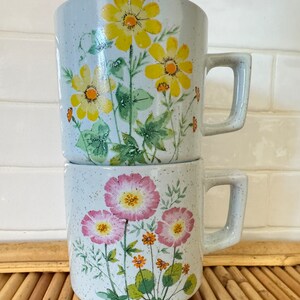 May include: Two vintage ceramic mugs with floral designs. The top mug has yellow flowers on a light gray background. The bottom mug has pink flowers on a light gray background.