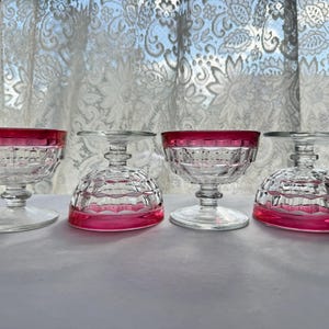 May include: Four clear glass dessert dishes with a pink rim. The dishes have a cut glass design and are arranged in a row on a white surface.