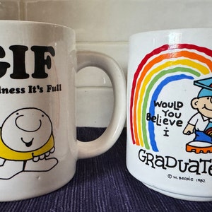 May include: Two white ceramic mugs with cartoon illustrations. The first mug has the text "TGIF Thank Goodness It's Full" and a cartoon character holding a mug. The second mug has a rainbow and the text "Would You Believe I Graduated?" with a cartoon character wearing a graduation cap.