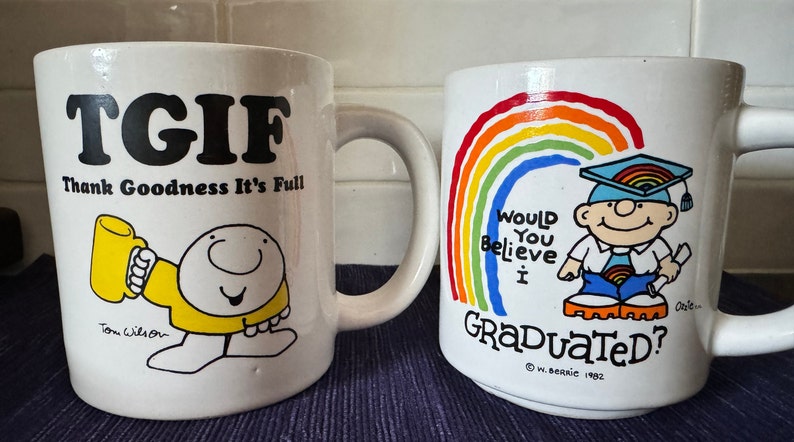 May include: Two white ceramic mugs with cartoon illustrations. The first mug has the text "TGIF Thank Goodness It's Full" and a cartoon character holding a mug. The second mug has a rainbow and the text "Would You Believe I Graduated?" with a cartoon character wearing a graduation cap.