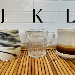 May include: Three mugs on a bamboo surface. The mug on the left is white with black marbling. The mug in the center is clear glass with a textured design. The mug on the right is white with brown stripes.