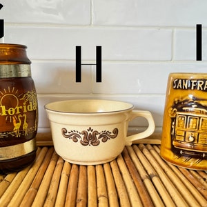 May include: Three ceramic mugs with different designs. The first mug is brown with a gold band and says "Florida" on it. The second mug is cream colored with a brown floral design. The third mug is brown with a picture of a San Francisco cable car and says "San Francisco" on it.