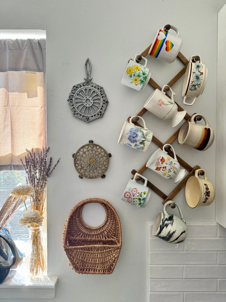 May include: A wooden mug rack with a variety of colorful mugs with floral and geometric designs. The rack is mounted on a white wall with a wicker basket and two trivets.