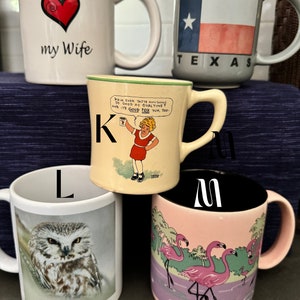 May include: A set of six white ceramic mugs with different designs. The mugs are labeled with the letters I, J, K, L, M, and W. The mug labeled I has a red heart with the words "I love my wife". The mug labeled J has a blue and red Texas flag with the words "Corpus Christi, Texas". The mug labeled K has a cartoon girl holding a can of Ovaltine with the words "Did ja ever taste anything so good as Ovaltine? And it's good for yum, too". The mug labeled L has a picture of an owl. The mug labeled M has a picture of pink flamingos. The mug labeled W is blank.