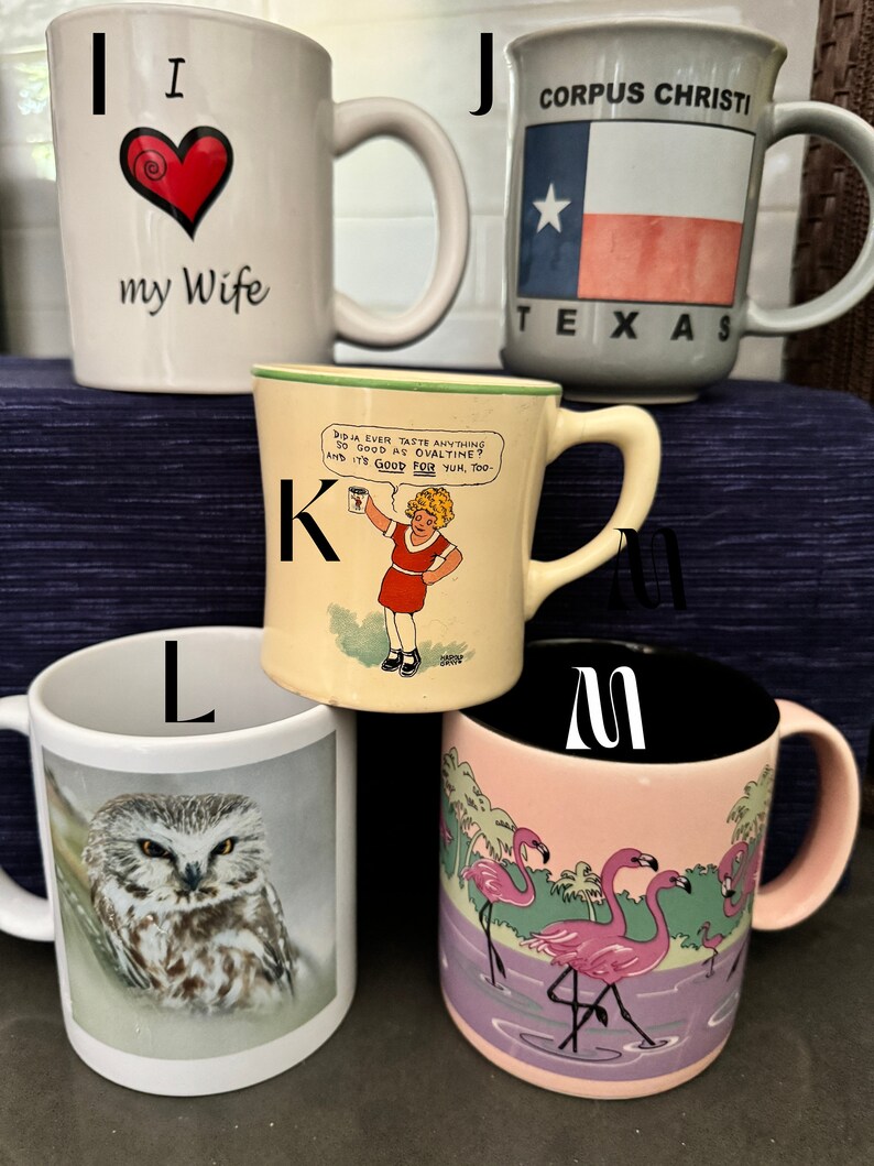 May include: A set of six white ceramic mugs with different designs. The mugs are labeled with the letters I, J, K, L, M, and W. The mug labeled I has a red heart with the words "I love my wife". The mug labeled J has a blue and red Texas flag with the words "Corpus Christi, Texas". The mug labeled K has a cartoon girl holding a can of Ovaltine with the words "Did ja ever taste anything so good as Ovaltine? And it's good for yum, too". The mug labeled L has a picture of an owl. The mug labeled M has a picture of pink flamingos. The mug labeled W is blank.