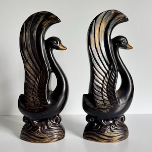 May include: A pair of black swan figurines with gold-toned highlights on the wings and base. Each swan is depicted with an arched neck and raised wings, standing on a textured, wave-like base. The finish is dark with metallic gold accents.