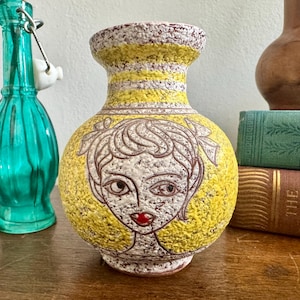 May include: Yellow ceramic vase with a white and brown stylized portrait of a person with red lips. The vase has a textured surface.