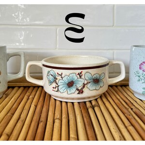 May include: Three ceramic mugs with floral designs. The mugs are white with speckles and feature different floral patterns in yellow, blue, and pink.