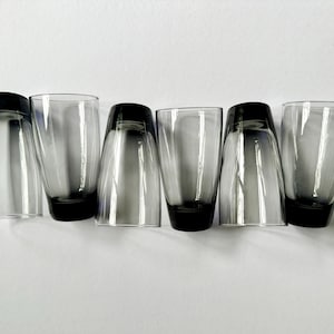 May include: Six clear shot glasses with a dark gray base and rim. The glasses are arranged in a row, with some upright and some inverted. The shot glasses are made of glass and are on a white background.