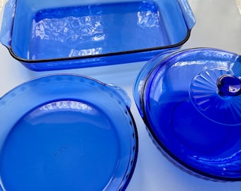 May include: Three pieces of blue glass bakeware. The set includes a rectangular baking dish with black trim, a round pie dish, and a round casserole dish with a lid. The dishes are a vibrant shade of blue.