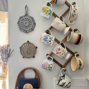 May include: A wooden mug rack with a variety of mugs hanging on it. The mugs are decorated with floral patterns, rainbows, and other designs. There are also two trivets on the wall, one made of metal and one made of wood.