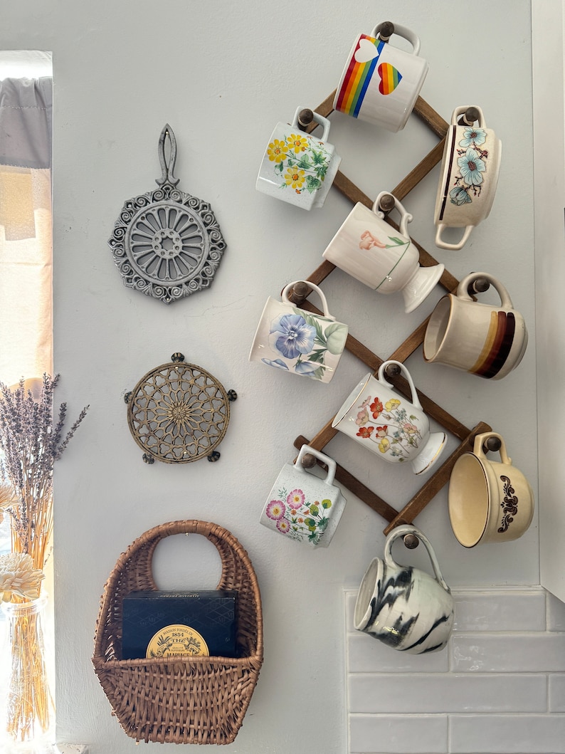 May include: A wooden mug rack with a variety of mugs hanging on it. The mugs are decorated with floral patterns, rainbows, and other designs. There are also two trivets on the wall, one made of metal and one made of wood.