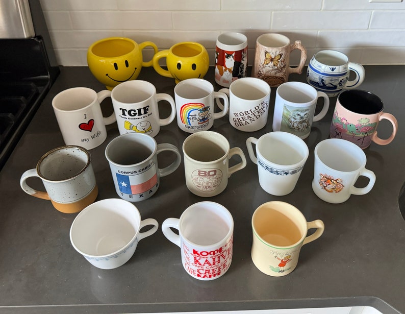 May include: A collection of 18 vintage ceramic mugs with various designs and colors. The mugs feature a variety of themes, including  "I love my wife", "TGIF", "World's Greatest Grandpa", "Corpus Christi, Texas", and "Kopi Kafe".