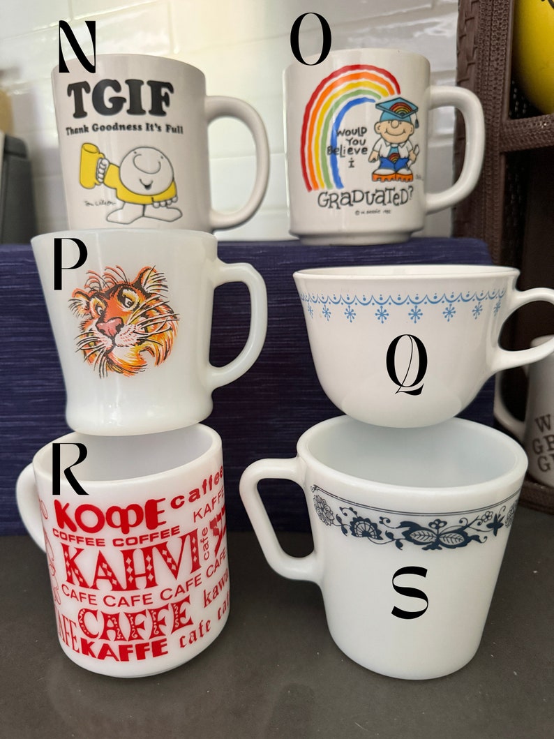 May include: Five white ceramic mugs with different designs. The mugs are arranged in a circle. The mugs have the following designs: "TGIF Thank Goodness It's Full", a rainbow with the text "Would You Believe I Graduated?", a tiger, a blue and white floral pattern, and a red and white pattern with the text "Kaffee, Cafe, Kopi, Kahvi".