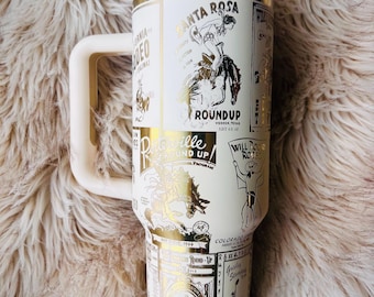 Peut inclure: Une tasse de voyage blanche et dorée avec une poignée. La tasse présente un design vintage avec des images de cowboys et d'événements de rodéo. Le texte sur la tasse comprend "Santa Rosa Roundup" et "Datepille Round Up".