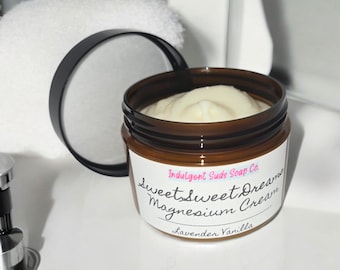 May include: A brown glass jar with a white label that reads "Indulgent Suds Soap Co. Sweet Sweet Dreams Magnesium Cream Lavender Vanilla". The jar is filled with a white cream.