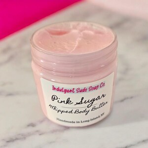 May include: A pink jar of whipped body butter with a white label that reads "Indulgent Suds Soap Co. Pink Sugar Whipped Body Butter Handmade in Long Island NY".