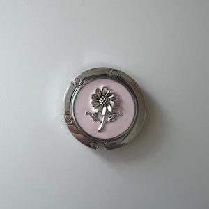 May include: A silver-toned purse hook with a pink center. The center features a detailed silver flower design. The hook is round and has a hinged design for easy storage and use. A stylish and functional accessory.