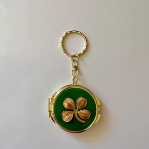 May include: A round gold-tone metal keychain featuring a hinged locket design. The front surface is inlaid with a smooth, deep green enamel finish and centered with a raised, textured gold-tone four-leaf clover emblem attached to a metal ring.