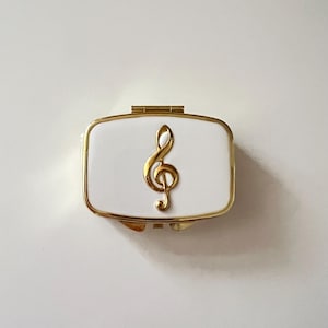 May include: A rectangular white pillbox with a gold treble clef emblem. The pillbox has a gold trim and a hinged lid. The design is simple and elegant, suitable for storing small items. The overall aesthetic is classic and refined.