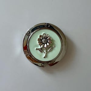 May include: A silver-toned purse hook with a light green center. A detailed flower design is in the center of the hook. The hook is round and has a hinged design for easy storage and use. The hook is designed to hang a purse from a table.
