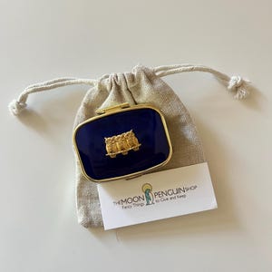 May include: A navy blue rectangular compact with a gold-colored frame and a gold owl design. The compact is inside a beige drawstring bag, with a white business card that reads "THE MOON PENGUIN SHOP Fancy Things to Give and Keep."