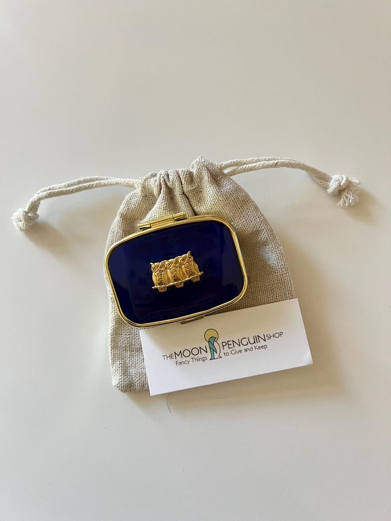 May include: A navy blue rectangular compact with a gold-colored frame and a gold owl design. The compact is inside a beige drawstring bag, with a white business card that reads "THE MOON PENGUIN SHOP Fancy Things to Give and Keep."