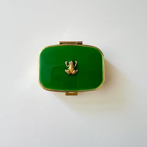 May include: A rectangular hinged box with a glossy emerald green lid and a polished gold-tone metal frame. A small, three-dimensional gold-colored frog figurine is centered on the top surface of the lid, facing forward with its legs extended.
