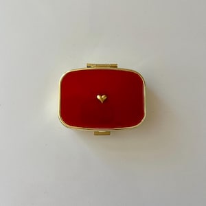 May include: A small, rectangular, red compact with a gold-colored frame. A tiny gold heart is centered on the red surface. The compact is closed, with a gold hinge visible.