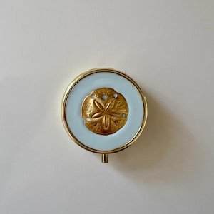 May include: A round gold-tone compact mirror featuring a light blue enamel inlay on the lid. Centered on the light blue surface is a raised, textured gold-tone sand dollar emblem with five distinct petal-like shapes and small circular perforations.