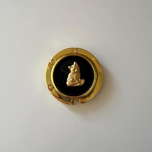May include: A circular gold-tone metal bag hanger featuring a black circular inlay in the center. A small, three-dimensional gold-colored cat figure is attached to the middle of the black inlay. The hanger has a hinged design with visible rivets.