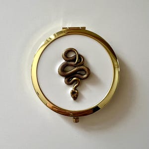 以下が含まれることがあります： 白の内側を持つ丸い金色のコンパクトミラー。詳細なブロンズ色のヘビが白い表面の中央に配置されています。コンパクトはヒンジ開口部と光沢のある反射仕上げです。