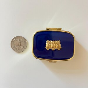 May include: A rectangular, navy blue pillbox with a gold-colored frame and hinge. The lid features a gold-colored relief of three owls. A U.S. quarter coin is next to the pillbox for size comparison.