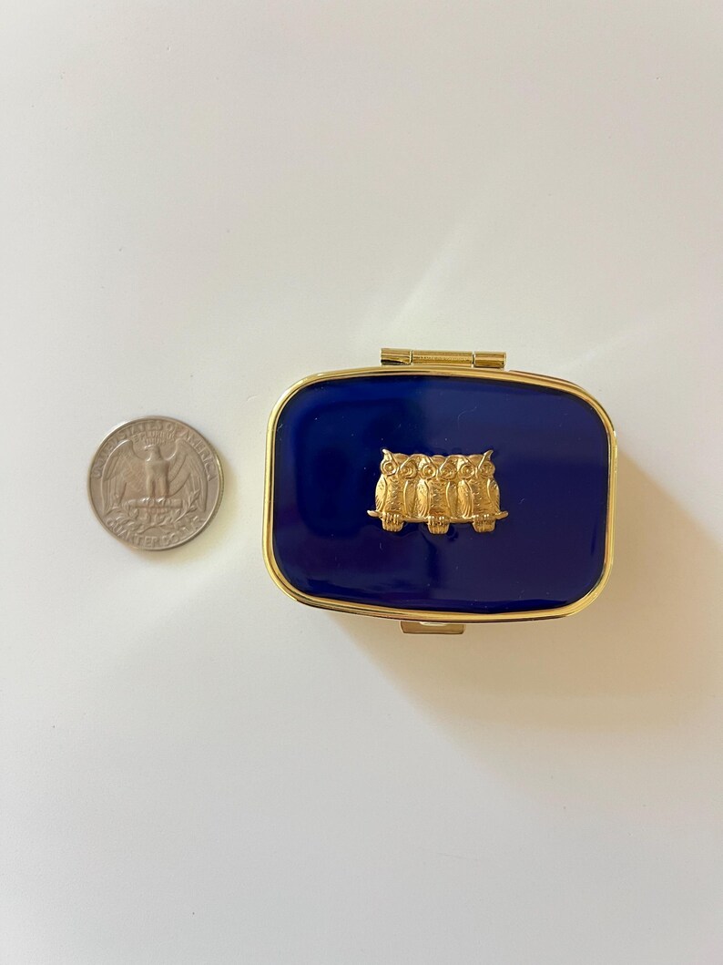 May include: A rectangular, navy blue pillbox with a gold-colored frame and hinge. The lid features a gold-colored relief of three owls. A U.S. quarter coin is next to the pillbox for size comparison.