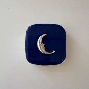 May include: A navy blue compact with a silver crescent moon design. The compact is square with rounded corners and a glossy finish. The moon has a detailed face.