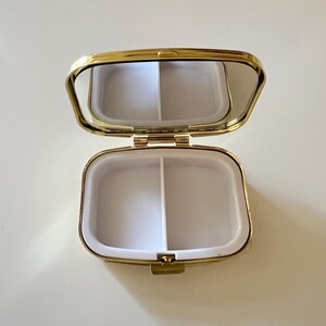 May include: Open, gold-toned pill box with a mirrored lid. The interior is divided into two white compartments. The pill box is rectangular with rounded edges and a clasp closure.